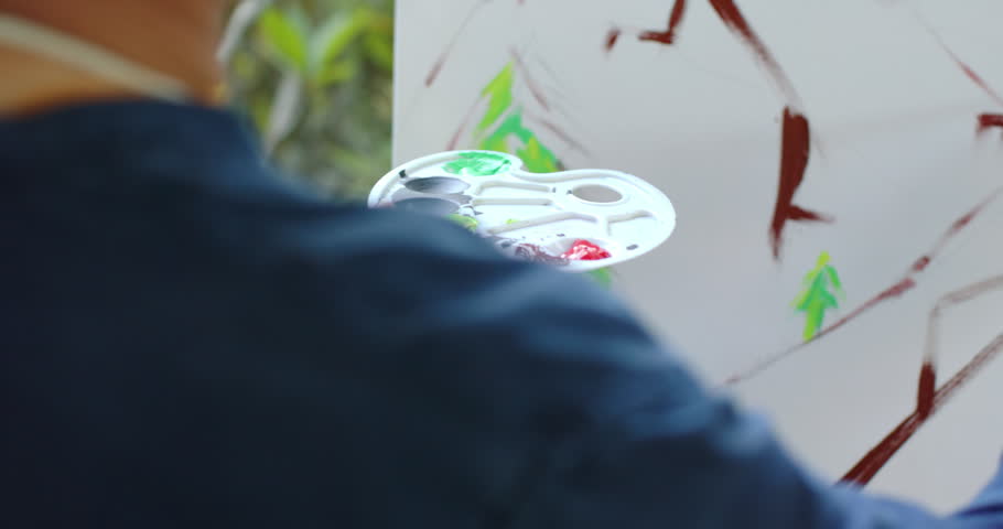 Senior Asian painter mixing paint blocking ridges adding firs for view on garden canvas, copy space. Artist, palette, brush, easel, landscape, mountains, trees