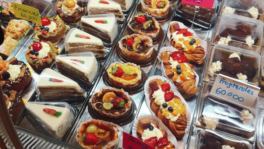 Bakery display showcasing a wide assortment of pastries, cakes, carrot cake slices, fruit tarts, croissants with cream and berries, and chocolate desserts, reflecting variety and tempting treats