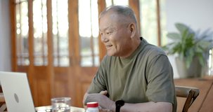 Senior Asian man sitting at dining table leaning to laptop speaking gesturing after incoming call. Home, daylight, wooden, plant, computer, conversation, engagement - Powered by Shutterstock - Get 15% off with code: PIKWIZARD15
