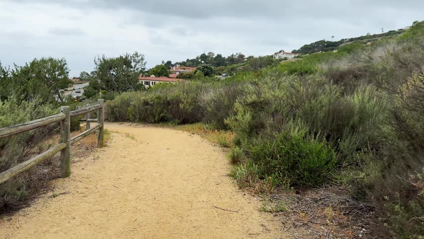 Unpaved hiking route, earthy tone, stretches through dense scrubland. Left side showcases weathered split rail fence. Right side features varied, drought tolerant plant life. Elevated background revea
