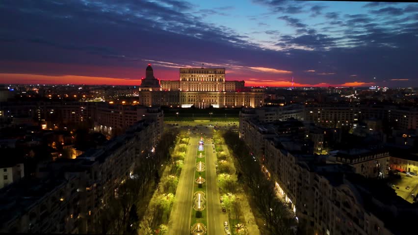 Cinematic aerial shot of the Palace of Parliament (Casa Poporului) in Bucharest, Romania. The camera moves forward and rises (pedestal up) during a dramatic sunset, showing the illuminated facade, city lights, and the vast urban landscape of the capital city.