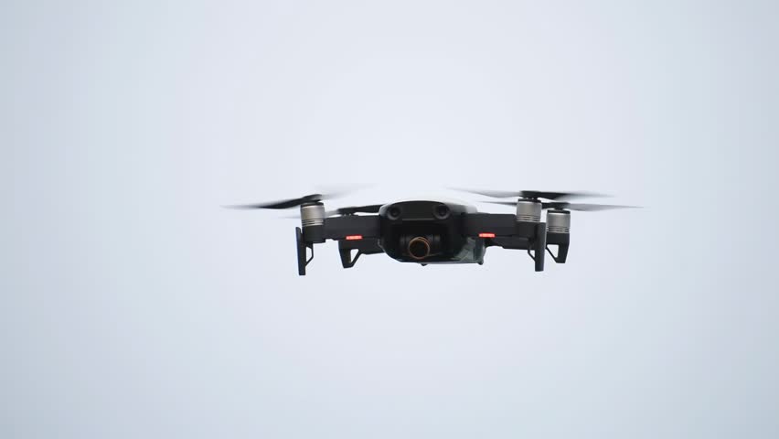 Modern quadcopter drone hovering in gray sky with spinning propellers and glowing red signal lights.