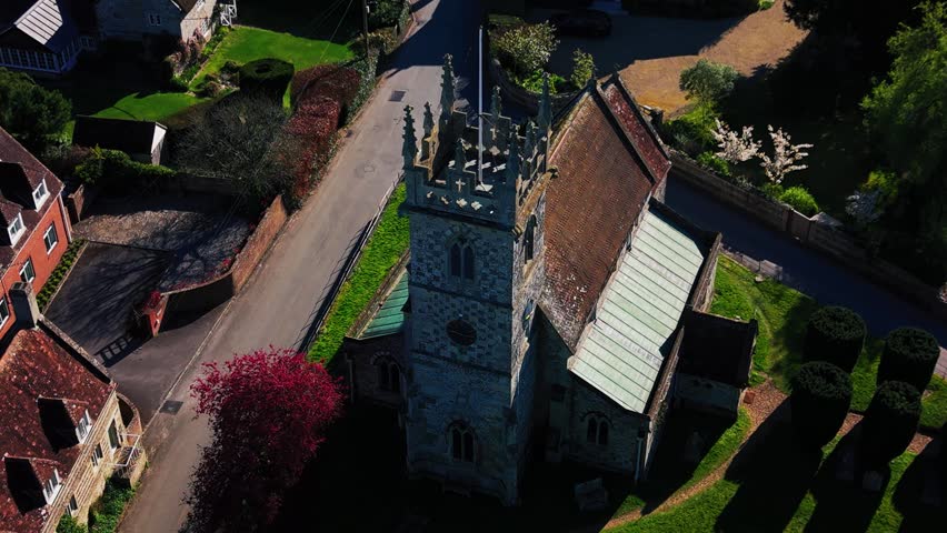 A close-up, high-angle aerial view of a historic stone church situated in a quiet English village. The shot heavily features the intricate architecture of the square church tower