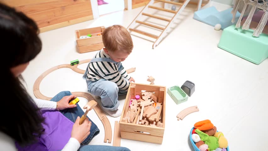 Over the shoulder view of a young boy and a daycare teacher sitting on the floor, collaborating to build a railway with a wooden train set, fostering early childhood development and learning