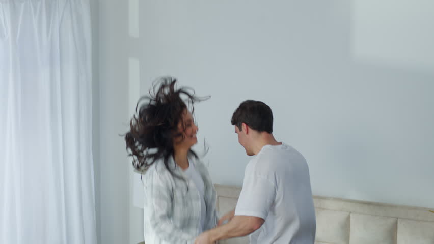 Happy Caucasian couple wife husband man woman jump on bed bedroom apartment fun boyfriend girlfriend jumping holding hands enjoyment laugh female male active bounce pampering move energy home together