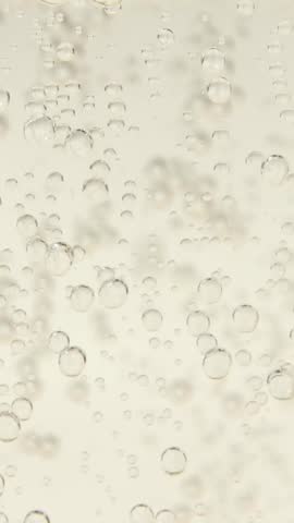 Macro closeup of a clear sparkling beverage as scattered bubbles appear across the frame, rise steadily upward, multiply through the liquid, and create a clean fizzy texture.