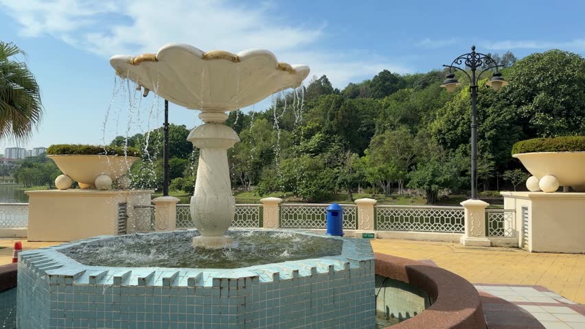 Water cascades from a white, shell-shaped fountain in a sunlit park. The scene features turquoise tiles, lush green hills in the background, and a serene lakeside walkway under a bright, clear sky.