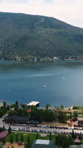 Vertical drone shot over Grand Lake Colorado showing the waterfront marina, broad mountain lake, evergreen shoreline, and alpine ridges stretching across the high country horizon