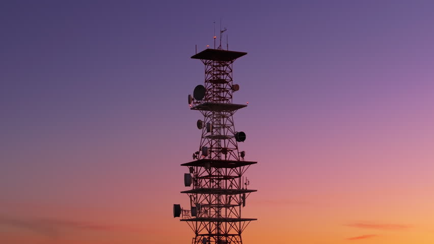 Tall telecommunications tower silhouetted against a stunning gradient sunset sky transitioning from purple to orange. Multiple satellite dishes and antennas with blinking red warning lights visible.