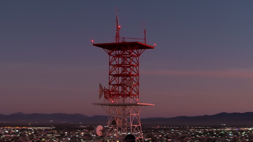 Aerial drone footage orbiting a red and white telecommunications tower with blinking aviation warning lights and microwave dishes against a purple twilight sky, city lights sparkling below.