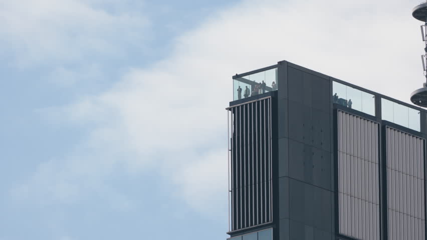 Ground level view looking up at modern skyscraper rooftop observation deck, people behind glass enjoying city views and taking photos, tower top against cloudy sky, cinematic urban perspective