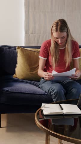 Vertical video: Woman on sofa sorting work papers for filing, mid circles appearing, marking pages. Documents, notebook, home, livingroom, organization, focus, vertical video
