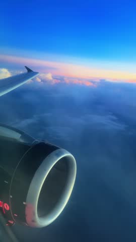 Vertical Screen: Airplane wing and jet engine visible from passenger window seat during flight above fluffy clouds at sunset with beautiful pink and orange gradient sky on the horizon.
