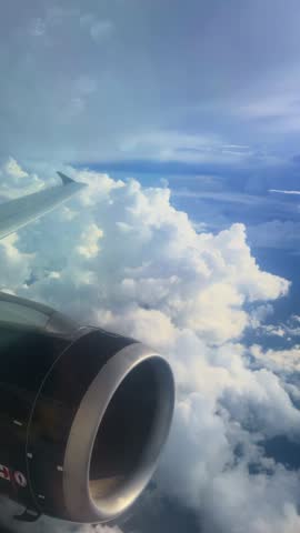Vertical Screen: Airplane jet engine and wing soar high above dramatic white cumulus clouds against a vibrant blue sky, with sunlight streaming through the cabin window creating soft lens flares.
