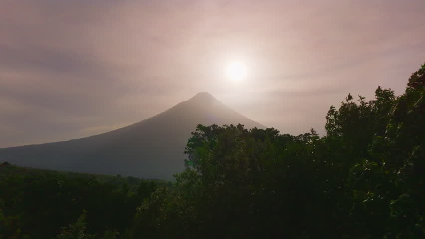 Majestic Volcano Arenal rises above lush Costa Rica rainforest during golden sunrise. Soft morning light illuminates the conical peak while mist drifts across dense jungle canopy below.
