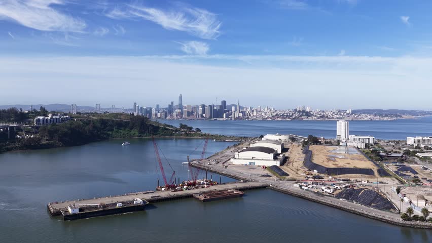 Aerial view flying over a large construction site on treasure island with the iconic san francisco city skyline and bay bridge visible in the background under a sunny, blue sky