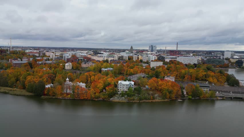 A stunning aerial view of Pyynikki Ridge and Lake Pyhäjärvi in Tampere, Finland, showcasing an autumn forest hillside blending into a tranquil lakeside cityscape, captured by drone in vivid colors.