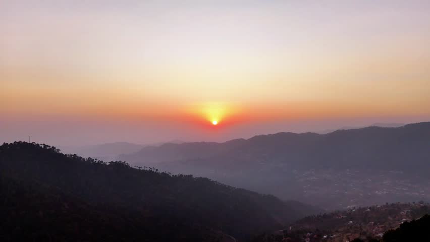 Stunning aerial drone view of a vibrant orange sunset over the silhouettes of the Margalla Hills mountain range, featuring a soft hazy horizon and tranquil evening sky in Islamabad, Pakistan.