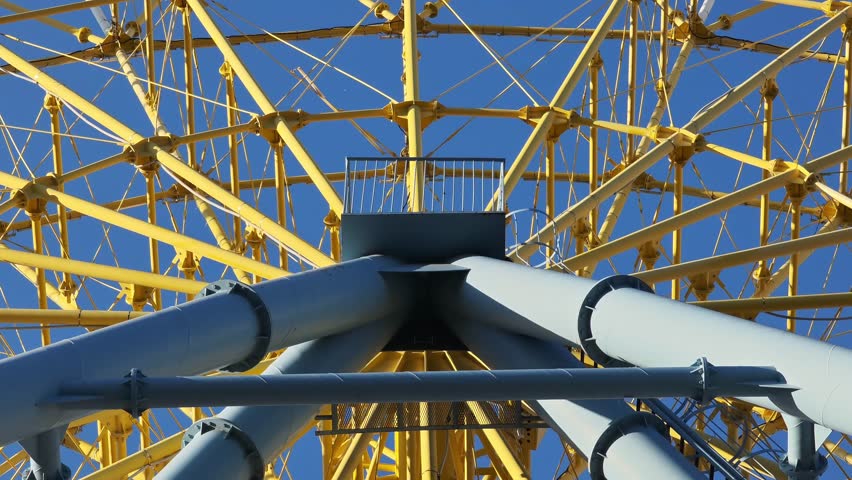 Close up of a giant yellow Ferris wheel spinning against a perfectly clear blue sky. The intricate steel structure slowly rotates