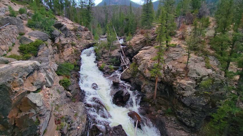 Waterfall scene resembling Rocky Mountain National Park in Colorado, with rushing whitewater flowing through rocky cliffs and pine forest in a rugged mountain landscape near Estes Park.