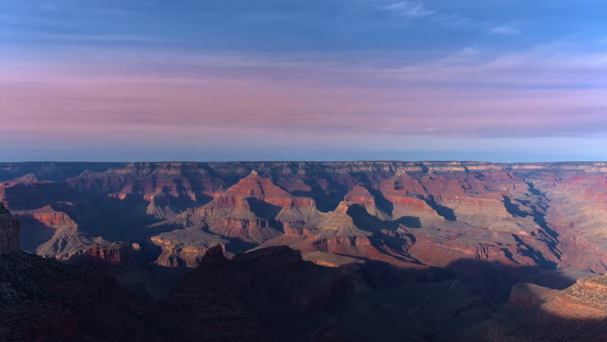 Arizona sunset time-lapse captures the Grand Canyon as golden hour light transitions across ancient layered rock formations. Pink and purple clouds drift over vast canyon depths during twilight.