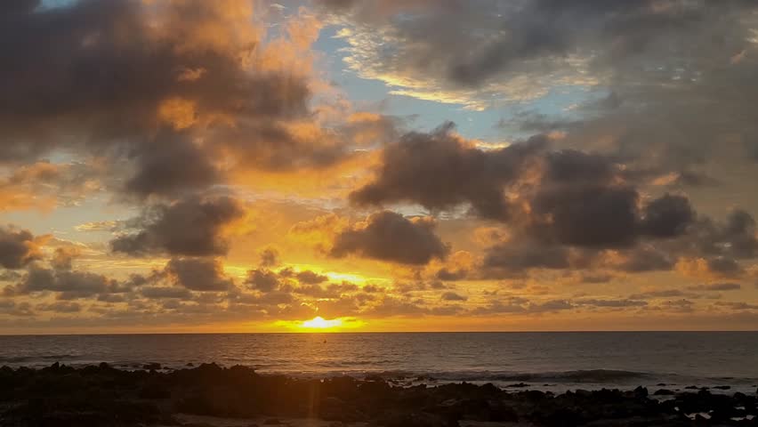 A breathtaking sunset over a vast ocean horizon. The sky is a dramatic canvas of deep blues and greys, contrasted by vibrant golden and orange hues radiating from the sun as it dips toward the water. Large, fluffy clouds are scattered across the frame, catching the warm light. In the foreground, a dark, rocky shoreline frames the view, while a tiny, solitary boat can be seen silhouetted against the shimmering reflection of the sun on the gentle waves