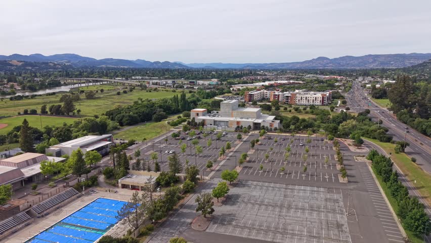 Expansive panning aerial view of Napa Valley College campus in Napa, California, showing mostly empty parking lot, covered swimming pool, green grounds and distant rolling mountains