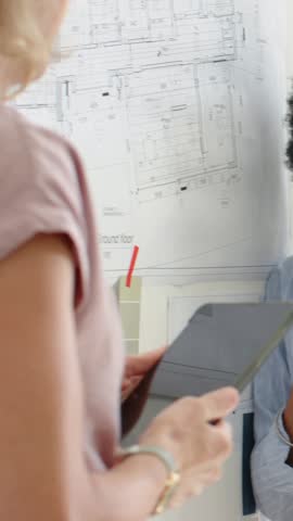 Vertical video: Listening woman in striped shirt responding to colleague presenting tablet at plans. Design, collaboration, blueprint, studio, architecture, eyewear, vertical video