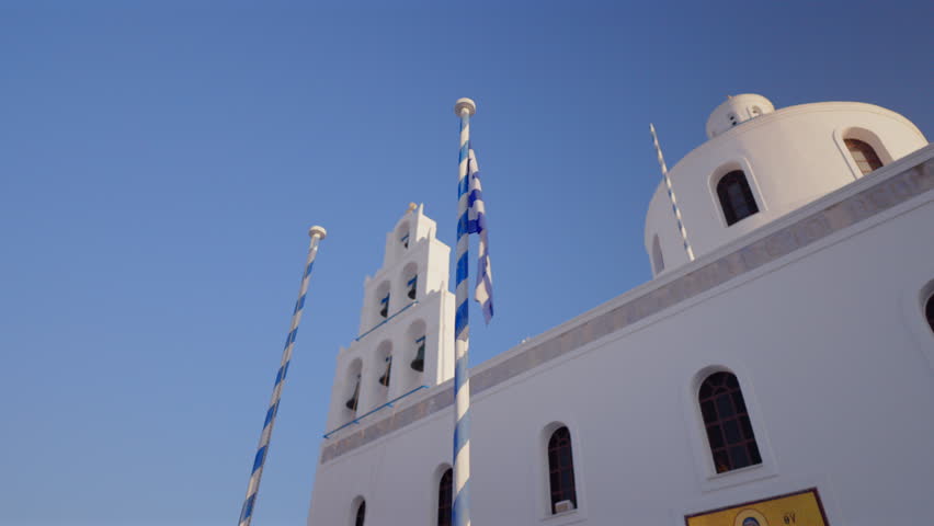 Greek national flag flutters on striped pole in front of traditional white Orthodox church featuring dome and bell tower. Clear blue sky frames iconic Cycladic architecture. Santorini, Greece.