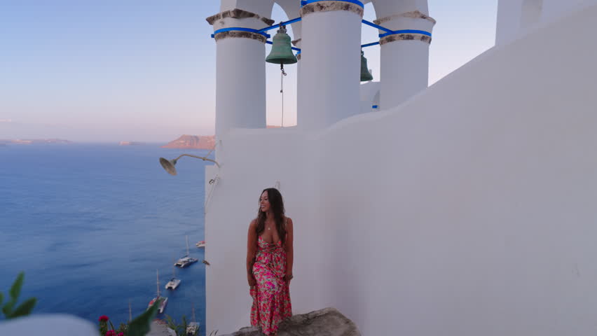 Beautiful woman wearing colorful floral maxi dress poses near traditional white Greek Orthodox church bell tower with scenic Aegean Sea and boats in background during golden hour. Santorini, Greece