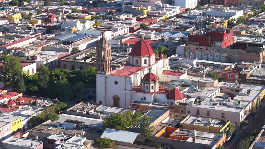 Aerial drone footage captures stunning colonial cathedral with distinctive red domes and baroque bell tower rising above the urban landscape of Durango Mexico. Green trees surround the historic.
