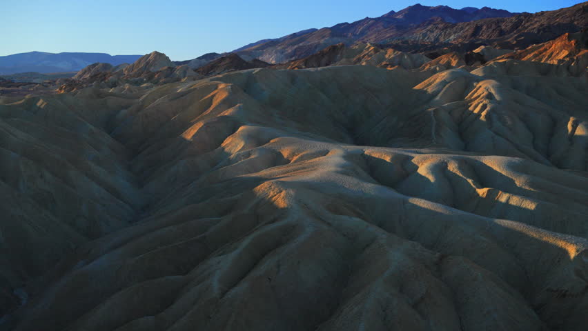 Dramatic eroded badlands landscape at Zabriskie Point in Death Valley National Park during early morning. Golden sunlight illuminates ridges of ancient sedimentary rock formations under clear blue.