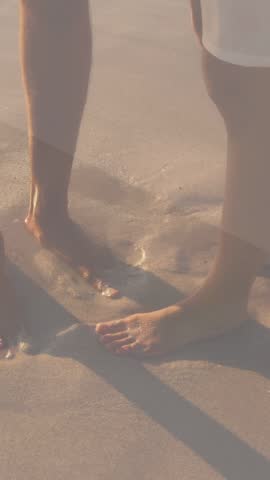 Vertical video: Pair standing in surf waves nudging toes tide overlay mapping water rise for travel. Couple, beach, shore, foam, footprints, goldenhour, vertical video