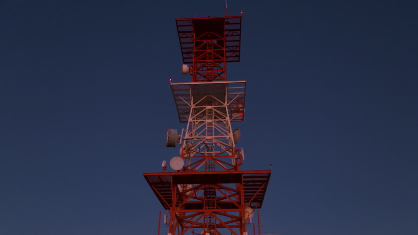 Aerial drone footage orbiting red and white telecommunications tower with satellite dishes and warning lights against a stunning gradient twilight sky over illuminated urban sprawl of Durango, Mexico.