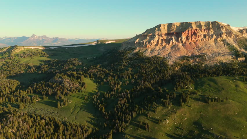 Beartooth Highway in Montana reveals stunning mountain landscape with red rock cliffs and lush green valleys. Pine forests stretch across rolling hills under golden evening light.