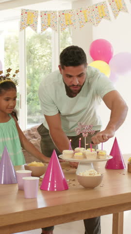 Vertical video: Diverse family having birthday dad lighting candles girl blowing and mom tossing hat. Celebration, family, joy, warmth, togetherness, colorful, vertical video