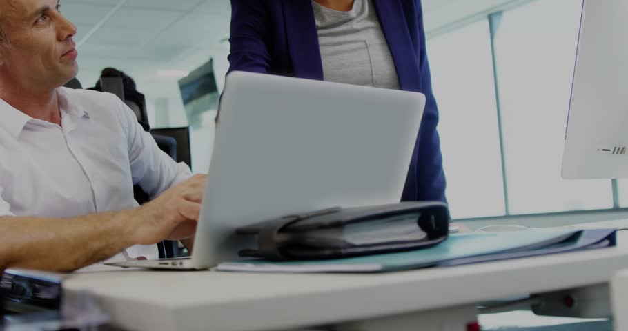 Seated man typing, looking, woman leaning in pointing at laptop with floating secure IT access tags. Collaboration, teamwork, office, monitor, binder, desk, encryption