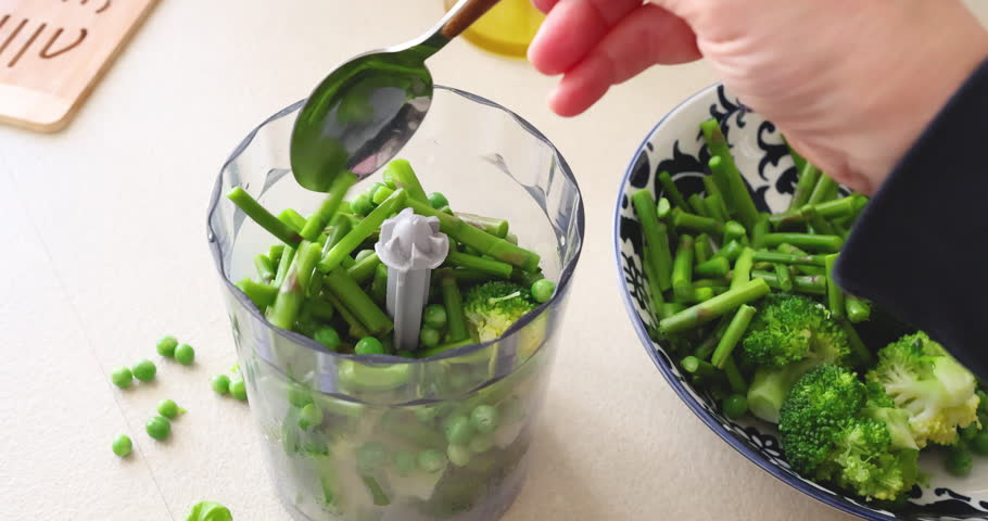 Fresh green vegetables like asparagus and broccoli with peas in blender container. Woman hand transferring organic ingredients from ceramic bowl for making healthy puree or soup. Culinary process of