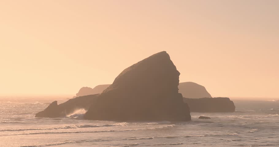 Silhouetted sea stacks standing in the ocean during a hazy sunset with gentle waves washing ashore in Oregon.