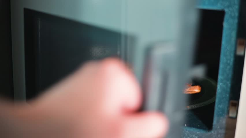 A hand reaches into a microwave to place a slice of leftover pizza on a plate. The pizza is then removed after heating, showing a quick and simple meal preparation at home.