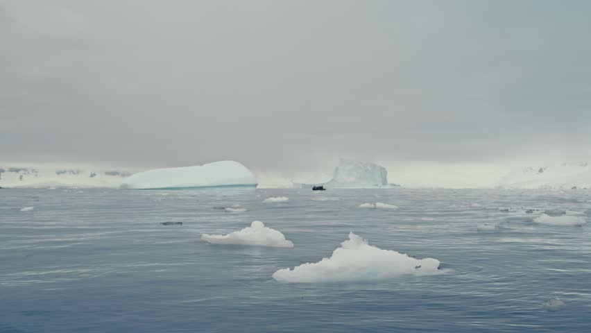 Small inflatable zodiac boat with tourists navigating among icebergs and floating ice in Antarctica under overcast sky