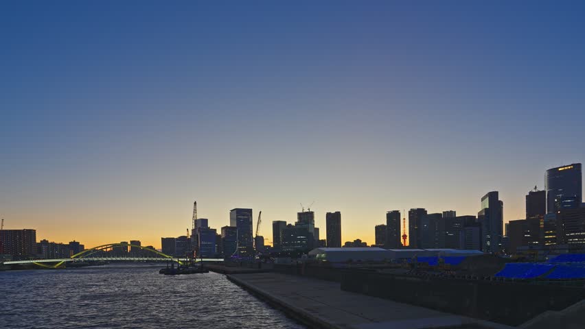 Tokyo waterfront skyline silhouettes against a warm orange-to-blue dusk sky, with a green arch bridge, barge on calm river, construction cranes, and a red tower visible, captured as a static wide shot