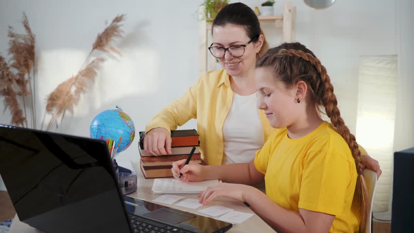 Mother helps daughter doing homework. Student sits writing. Mother points at laptop screen. Happy mother watches daughter studying. Cheerful daughter finishes homework. Laptop aids fast learning.