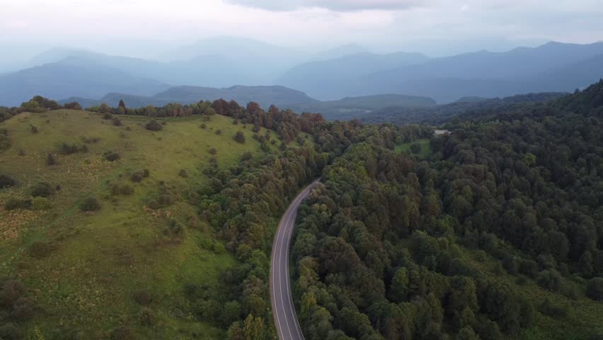 The winding road passes through green hills and dense forest landscape. The mountains stretch away into the distance. Sky with soft clouds, sunset.