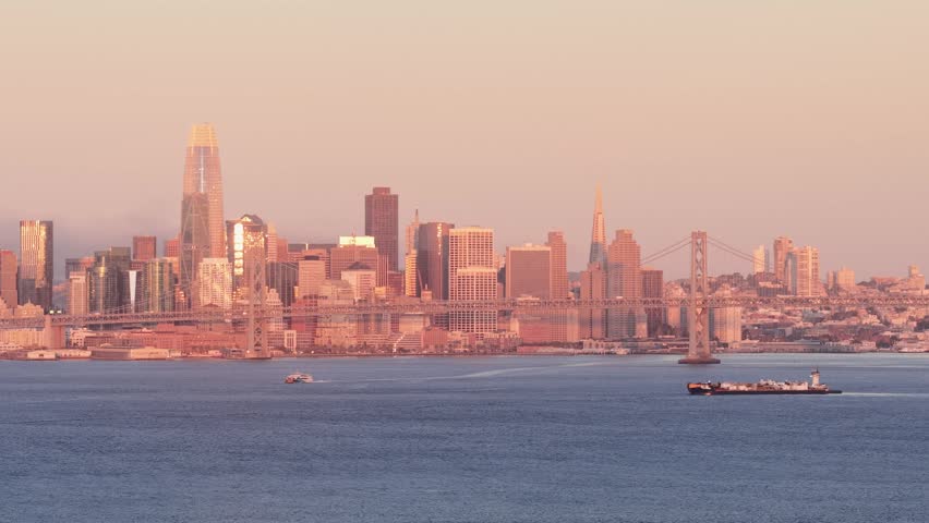 Sweeping view of the San Francisco skyline, including the Salesforce Tower and the Bay Bridge, bathed in the warm, golden light of sunrise as a cargo barge crosses the calm water