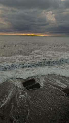 Stormy sunset over rocky shore, dark clouds with narrow golden horizon, storm chaser observation of crashing waves and foam