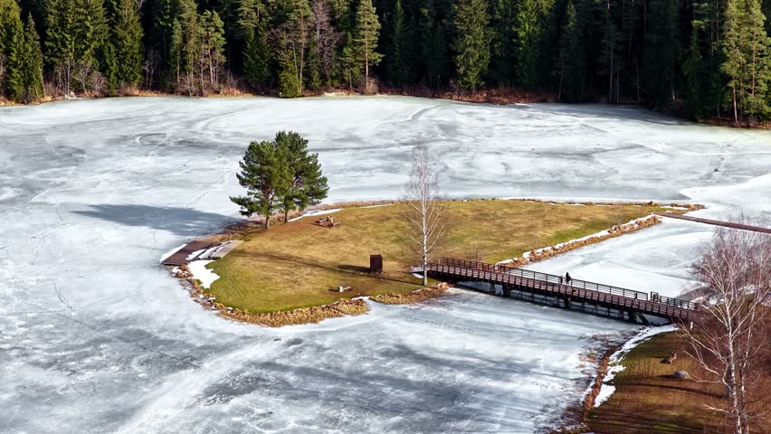 Winter lake island with walking bridge, frozen water and bare trees, aerial orbital shot