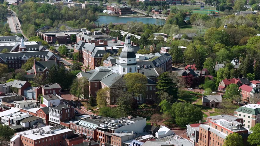 Aerial drone view of historic Annapolis, Maryland, USA with Maryland State House, old town streets, red brick buildings, calm river, bridge and lush spring cityscape in sunlight. Orbit shot.