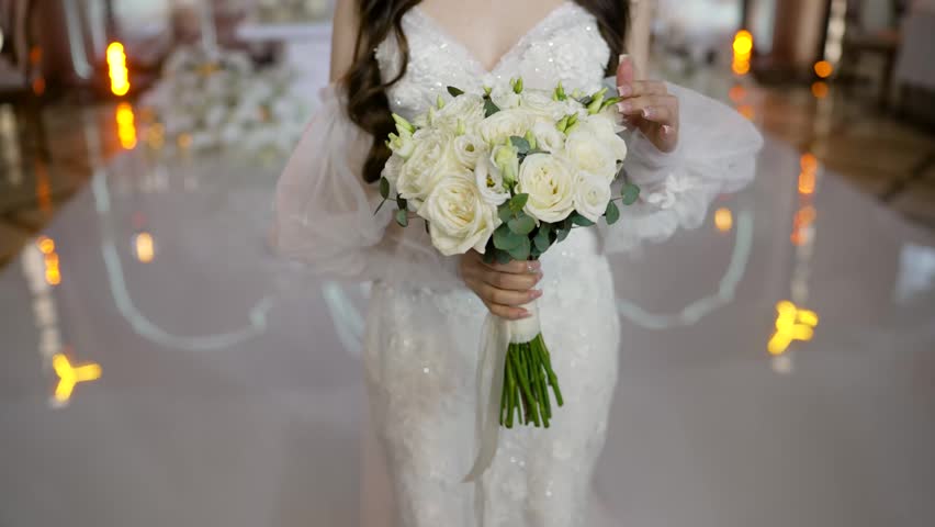 Bride in elegant white lace dress holds beautiful bouquet of white roses greenery. Close up shot focuses on flowers hands. Wedding day scene with soft lighting reflective floor background