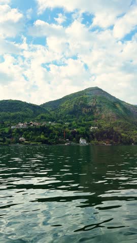 Beautiful view of Italian lakeside town with luxury villas nestled on green mountain slope under clouds at Lake Como during sunny spring day.